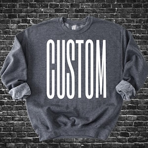 May include: A dark gray sweatshirt with the word "CUSTOM" printed in large, white, capital letters. The sleeves are cuffed, and the background is a dark brick wall.