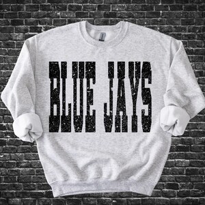 May include: A light grey sweatshirt featuring the text "BLUE JAYS" in large, distressed black lettering. The sweatshirt has a crew neck and rolled-up sleeves. The background is a dark brick wall.