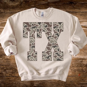 May include: A cream-colored sweatshirt with the letters "TX" in a camouflage pattern. The letters are in a block style, with the camouflage design featuring shades of brown, gray, and green.