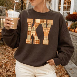 May include: A dark brown crewneck sweatshirt featuring the letters KY printed on the front. The letters are filled with a patchwork pattern of various plaid and checkered designs in shades of orange, yellow, cream, and muted green.