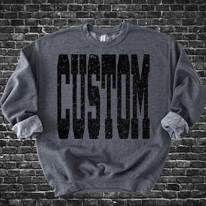 Pode incluir: Um moletom cinza escuro com a palavra "CUSTOM" impressa em letras pretas grandes e desgastadas. O moletom tem gola redonda e mangas enroladas. O fundo é uma parede de tijolos escuros.