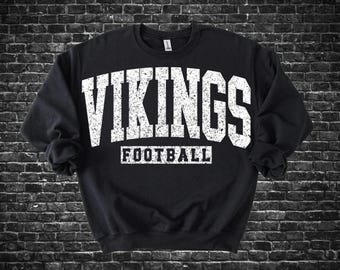 May include: Black crewneck sweatshirt with the word "VIKINGS" in large, distressed white letters across the chest, above the word "FOOTBALL" in a smaller, white, rectangular font. The sweatshirt is against a dark brick wall.