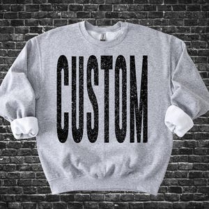 May include: A heather gray sweatshirt with the word "CUSTOM" printed in large, distressed black letters across the front. The sleeves are cuffed, revealing white fabric. The sweatshirt is displayed against a dark brick wall.