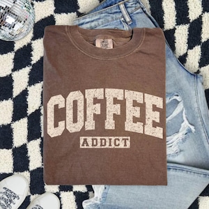 Może przedstawiać: Brązowa bluza z okrągłym dekoltem z napisem "COFFEE ADDICT" w postarzanym, złamanym białym kroju pisma. Bluza jest złożona na czarno-białym dywanie w szachownicę, z parą niebieskich dżinsów i białych tenisówek.