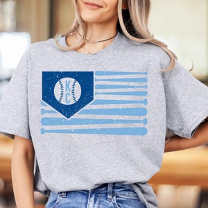 May include: A light gray t-shirt featuring a baseball-themed design. The design includes a blue flag with baseball bats and a baseball with the letters "KC". The shirt is made of a soft material.