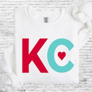 May include: White sweatshirt with a red "K" and a turquoise "C" with a red heart inside. The letters are large and bold.