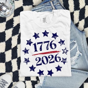 Pode incluir: Camiseta branca com os números "1776" e "2026" em azul marinho, rodeados por estrelas, com uma linha vermelha por baixo. A camiseta está sobre uma superfície xadrez em preto e branco com jeans e tênis brancos.