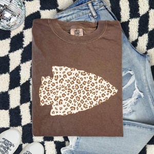 May include: A brown Comfort Colors crewneck sweatshirt featuring a leopard print arrowhead design. The sweatshirt is folded and rests on a black and white checkered rug, with a pair of blue jeans and white sneakers in the background.
