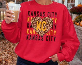 Może przedstawiać: Jaskrawoczerwona bluza z okrągłym dekoltem z powtarzającym się czarno-żółtym napisem KANSAS CITY. Centralny kształt serca z motywem panterki zawiera żółte litery KC. Wzór jest nadrukowany na przedniej części odzieży, tworząc wyraźny kontrast.