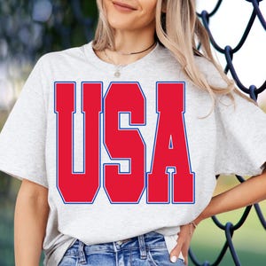May include: A light gray t-shirt with the word "USA" in large, bold, red letters with a blue outline. The shirt is a casual style, suitable for everyday wear.