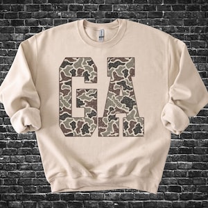 May include: A beige sweatshirt with the letters "GA" in a camouflage pattern. The letters are in a block style, and the sweatshirt has long sleeves with rolled cuffs. The background is a dark brick wall.