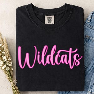 Op de afbeelding: Zwart T-shirt met het woord "Wildcats" in roze script. Het shirt is opgevouwen, naast een boeket gedroogde bloemen en een spijkerbroek.