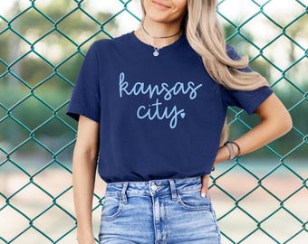 Może przedstawiać: Koszulka w kolorze granatowym z napisem "kansas city" w jasnoniebieskich literach z małym symbolem serca.