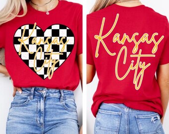 Może przedstawiać: Czerwona koszulka z czarno-białym sercem w kratkę z napisem "Kansas City" w złotym kolorze na przodzie. Z tyłu koszulka ma napis "Kansas City" w złotym kolorze.