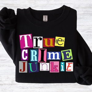 May include: Black sweatshirt with the text "True Crime Junkie" in a colorful, ransom-note style design. The text is composed of individual letters in various colors, including pink, blue, yellow, and green. The sweatshirt has long sleeves.