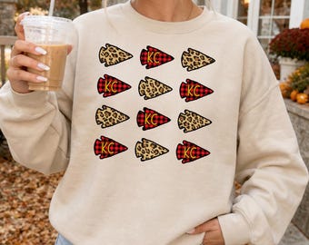 Op de afbeelding: Een beige sweater met ronde hals en een grafische print van twaalf pijlpuntsvormen in een raster. De pijlpunten wisselen af tussen een bruin luipaardpatroon en een rood met zwart ruitpatroon met de gele letters KC in het midden.