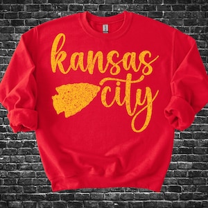 May include: A red sweatshirt with the words "Kansas City" in a yellow, cursive font. Below the words is a yellow arrowhead graphic. The sweatshirt has long sleeves and a crew neck.