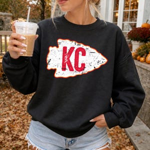 May include: A black long-sleeve sweatshirt featuring a white arrowhead graphic with a distressed texture and the red letters KC centered inside. The garment has a crew neckline and long sleeves with ribbed cuffs, providing a casual look.
