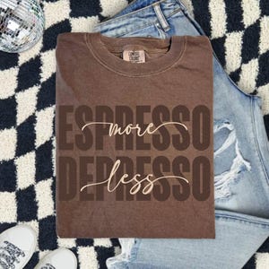May include: A brown Comfort Colors t-shirt with the slogan "Espresso More, Depresso Less" printed on the front in a mix of bold and cursive fonts. The shirt is folded on a black and white checkered rug, with jeans and white trainers.