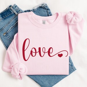 May include: A pink sweatshirt with the word "love" written in red cursive with a small red heart at the end.