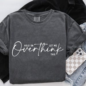 May include: Dark gray sweatshirt with the text "Hold on, let me overthink this" in white script. The sweatshirt is folded next to a pair of blue jeans and a phone with a checkered case.