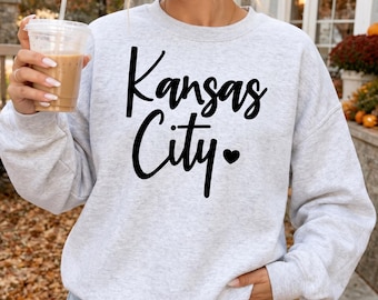 Może przedstawiać: Jasnoszara melanżowa bluza z okrągłym dekoltem, ozdobiona czarnym napisem Kansas City oraz małym symbolem serca. Odzież posiada długie rękawy oraz ściągacze przy dekolcie, mankietach i na dole bluzy.