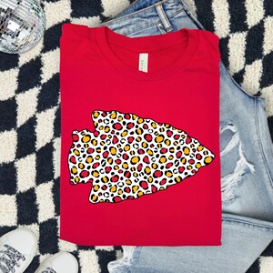 May include: A red t-shirt featuring a large arrowhead design filled with a leopard print pattern in red, yellow, and white. The shirt is folded and placed on a denim surface.