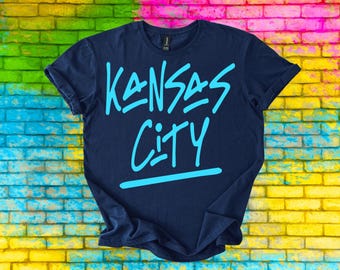 May include: Navy blue t-shirt with the words "Kansas City" in light blue, stylized lettering. The shirt is displayed against a colorful brick wall background.