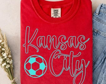May include: A red t-shirt with the words "Kansas City" in a teal script font, along with a teal soccer ball graphic. The shirt has a crew neck and short sleeves. The tag reads "Comfort Colors".