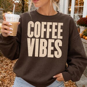 Pode incluir: Uma camisola de gola redonda castanho-escura com o texto COFFEE VIBES numa fonte grande, desgastada e de cor creme sem serifas no centro do peito. A peça tem mangas compridas e uma gola canelada, apresentando um visual simples e casual.