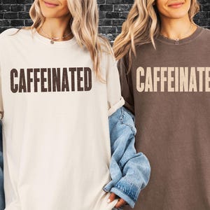May include: Two oversized t-shirts, one cream and the other brown, each displaying the word "CAFFEINATED" in a contrasting colour. The shirts have short sleeves and a relaxed fit, suitable for casual wear.