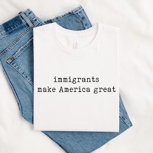 以下が含まれることがあります： 白いTシャツに、黒い文字で「immigrants make America great」とプリントされています。Tシャツは折り畳まれて、白い布の上に置かれ、青いジーンズが一緒に置かれています。