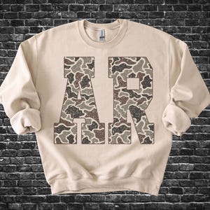 May include: A beige sweatshirt with the letters "AR" in a camouflage pattern. The letters are filled with a mix of brown, tan, and gray shapes. The sweatshirt has a crew neck and long sleeves with rolled cuffs.