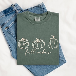Puede incluir: Una camiseta verde salvia con tres ilustraciones de calabazas y la frase "fall vibes" en cursiva. La camiseta está colocada sobre unos vaqueros azules. La camiseta es de la marca Comfort Colors.