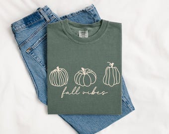 May include: A sage green t-shirt featuring three pumpkin designs and the words "fall vibes" in cursive. The t-shirt rests on blue denim jeans. The shirt is a Comfort Colors brand.