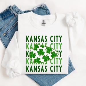 May include: White sweatshirt with the text "Kansas City" repeated four times in green and black. A green shamrock design in the shape of an arrow is between the words.