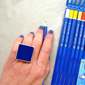 May include: A hand wearing a blue ring with a square blue stone, with blue nail polish, holding a blue pencil. The hand is in front of a box of 24 watercolor pencils, labeled "ergosoft" with the text "24 Buntstifte aquarell Coloured pencils aqua Crayons de couleur aquarellables" on the box.