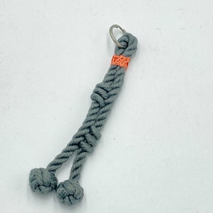 May include: A gray rope keychain with a knotted design. The keychain features a small orange accent near the top and two knotted balls at the bottom. A silver key ring is attached at the top.