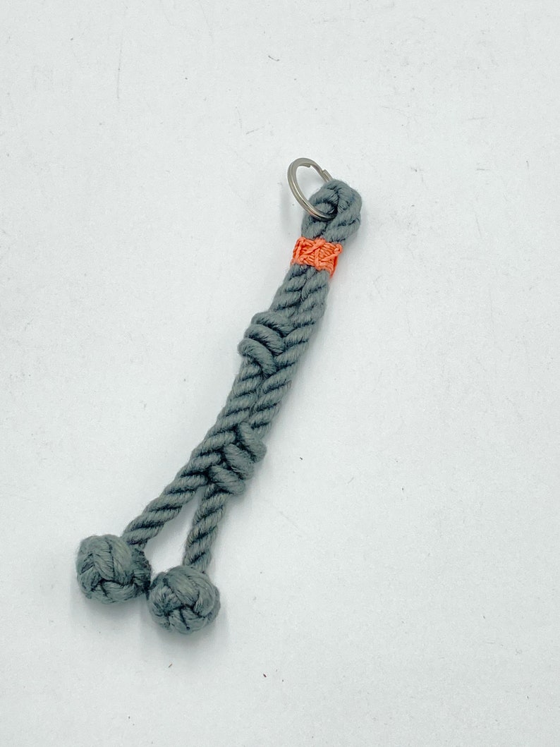 May include: A gray rope keychain with a knotted design. The keychain features a small orange accent near the top and two knotted balls at the bottom. A silver key ring is attached at the top.