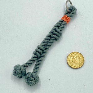 May include: A gray rope keychain with two decorative knots at the bottom. The keychain has a small orange accent near the top and a silver ring. A gold coin is next to the keychain.