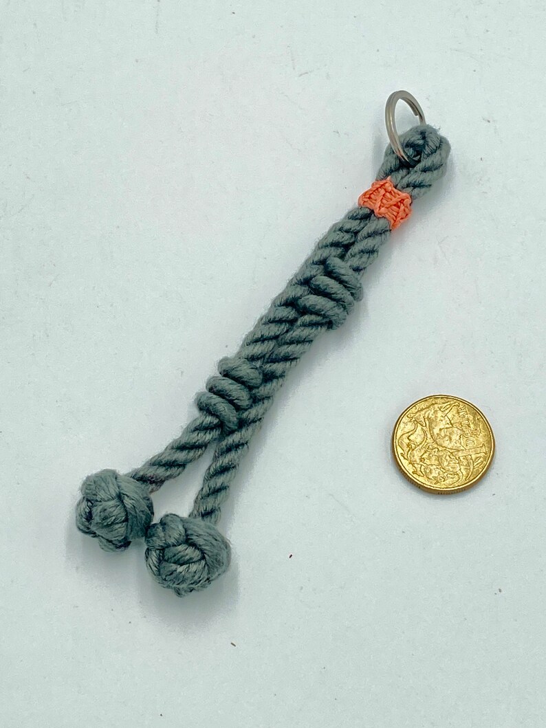 May include: A gray rope keychain with two decorative knots at the bottom. The keychain has a small orange accent near the top and a silver ring. A gold coin is next to the keychain.