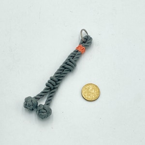 May include: A gray macrame keychain with two knotted ends. The keychain has a small orange accent and a silver key ring. A gold coin is next to the keychain.
