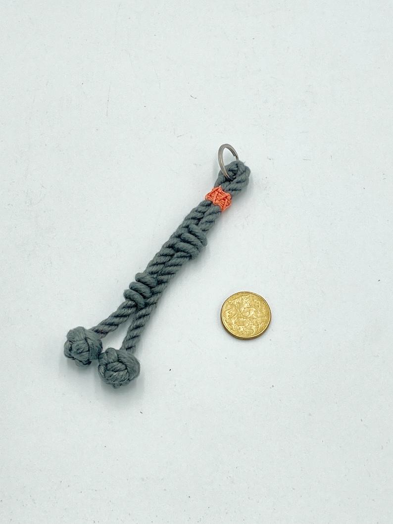 May include: A gray macrame keychain with two knotted ends. The keychain has a small orange accent and a silver key ring. A gold coin is next to the keychain.