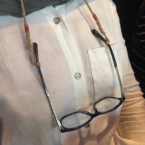 Könnte beinhalten: Eine Brille mit einem dunklen Gestell und klaren Gläsern hängt an einer beigefarbigen Schnurkette. Die Schnur hat rote Akzente an den Enden.