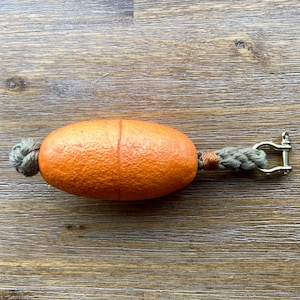 May include: Orange buoy with a textured surface, connected to a gray rope and a brass shackle. The buoy is oval-shaped and has a seam down the middle. The rope is knotted at one end.