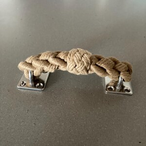Puede incluir: Un tirador de puerta decorativo con un diseño de cuerda trenzada en color beige natural. El tirador está sujeto a dos placas metálicas rectangulares plateadas con agujeros para tornillos. La cuerda está anudada en el centro.