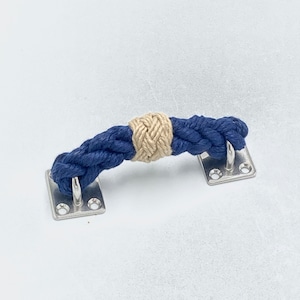 May include: A nautical-themed cabinet pull featuring a navy blue rope handle with a natural tan rope accent in the center. The handle is attached to two rectangular, silver-colored metal plates with screw holes.