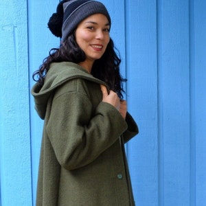 Könnte beinhalten: Eine Frau trägt einen dunkelgrünen Kapuzenmantel mit einer großen Tasche auf der linken Seite. Sie trägt eine schwarze Mütze mit einem Bommel oben und graue Stiefel.