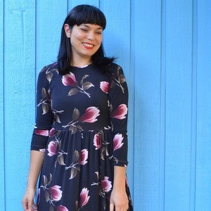 以下が含まれることがあります： 青い壁の前に立っている女性。黒い花柄のワンピースを着て、茶色のアンクルブーツを履いています。ワンピースは、体にフィットした上半身と、ゆったりとしたスカートになっています。