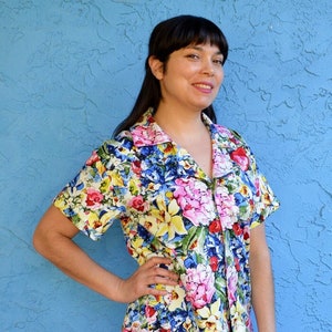 May include: A woman wearing a short-sleeved button-up shirt with a colorful floral print. The shirt has a collar and a button-up front. The woman is wearing black pants.
